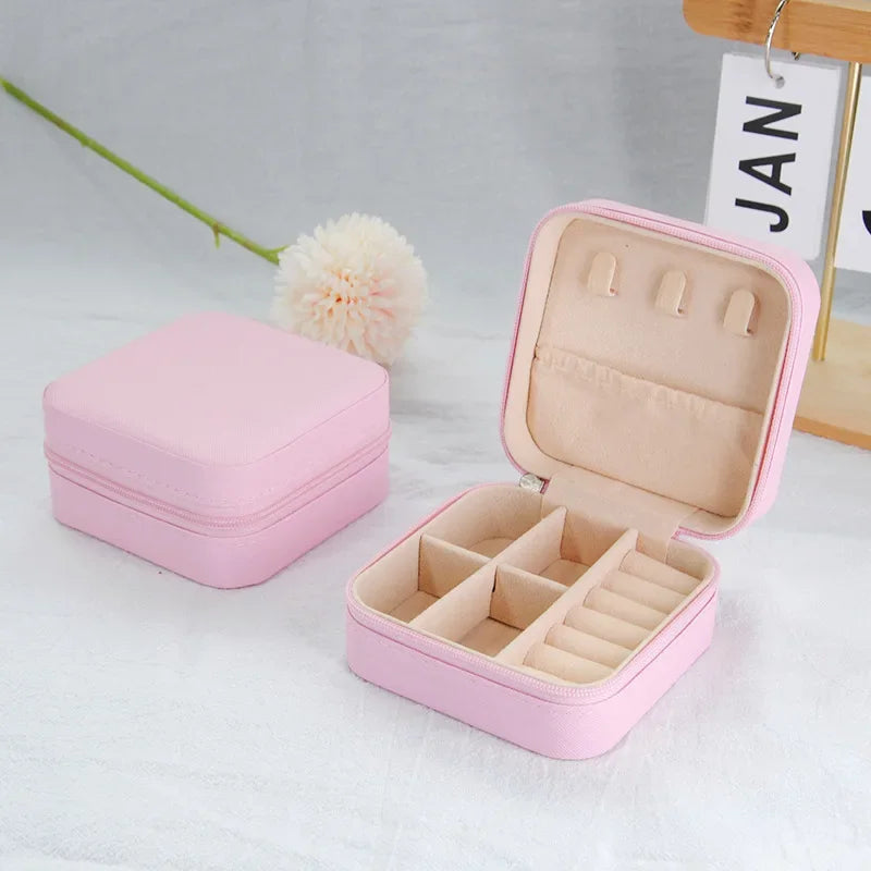 Two empty pink, square, zippered jewelry boxes, one open with organized compartments, on a white surface with a flower.