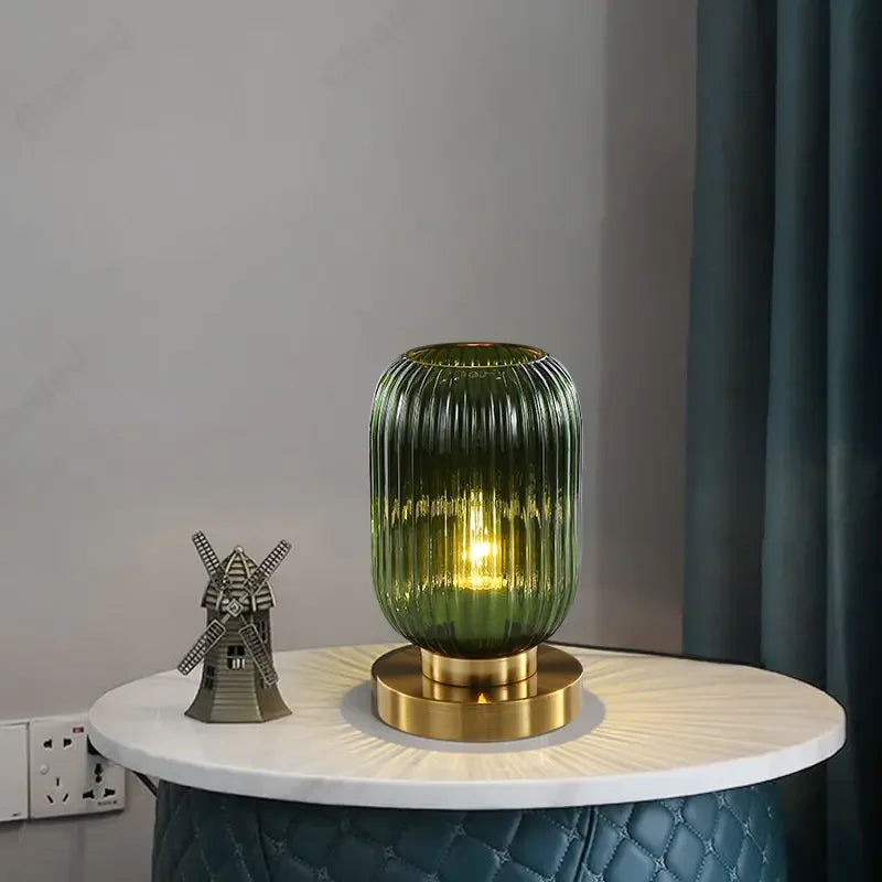 A lit, ribbed, green glass table lamp with a gold base sits on a small, round white table. A miniature windmill decoration is placed to its left, and a teal quilted surface is visible beneath the tabletop. A gray wall and a dark teal curtain form the backdrop.
