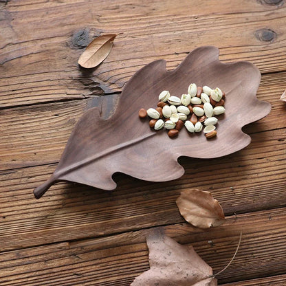 A wooden oak leaf-shaped platter holding pistachios and nuts, placed on a rustic wooden surface with dry leaves nearby.