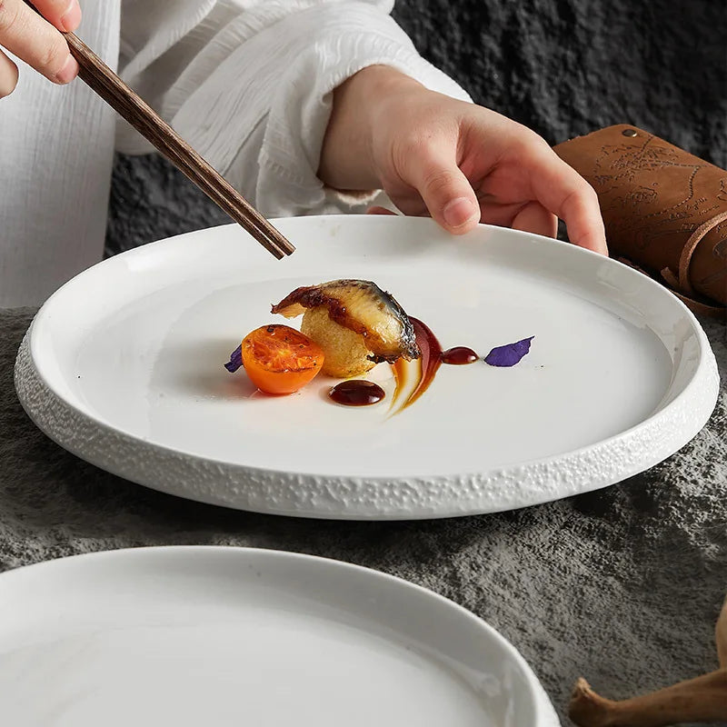 A minimalist white stoneware plate with a textured rim, elegantly plated with a gourmet dish, as a hand delicately uses chopsticks to pick up a bite.