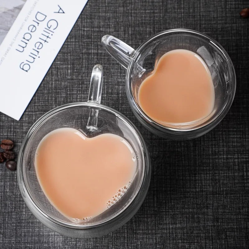 two heart-shaped glass mugs filled with coffee are on a dark surface, with a book and coffee beans nearby.