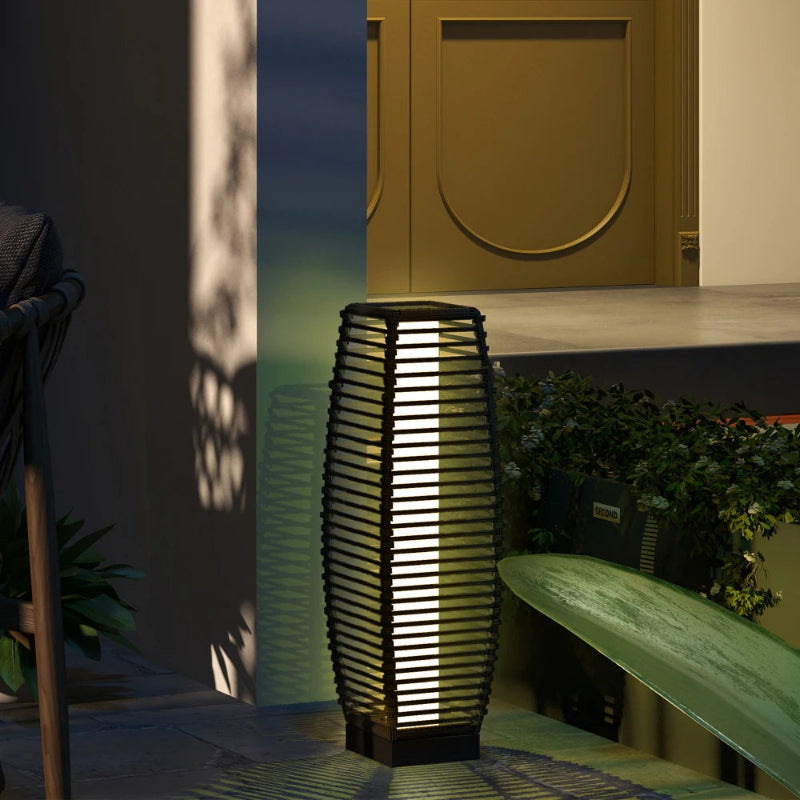 A tall, cylindrical outdoor lantern with a slatted black frame illuminates a patio at dusk.