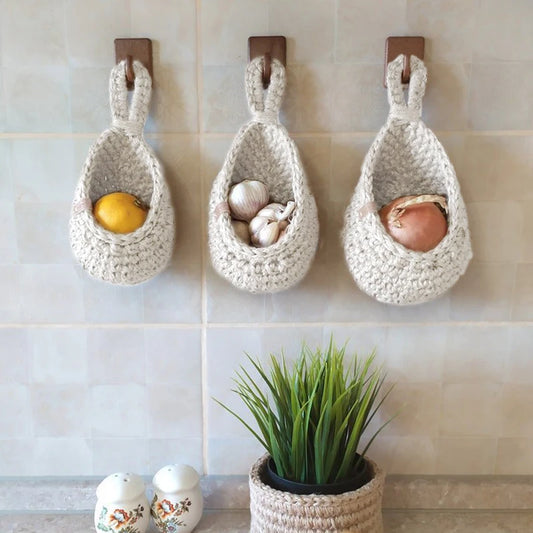 Three crocheted hanging baskets on a tiled wall, each holding a different food item, with a potted plant and salt and pepper shakers below.