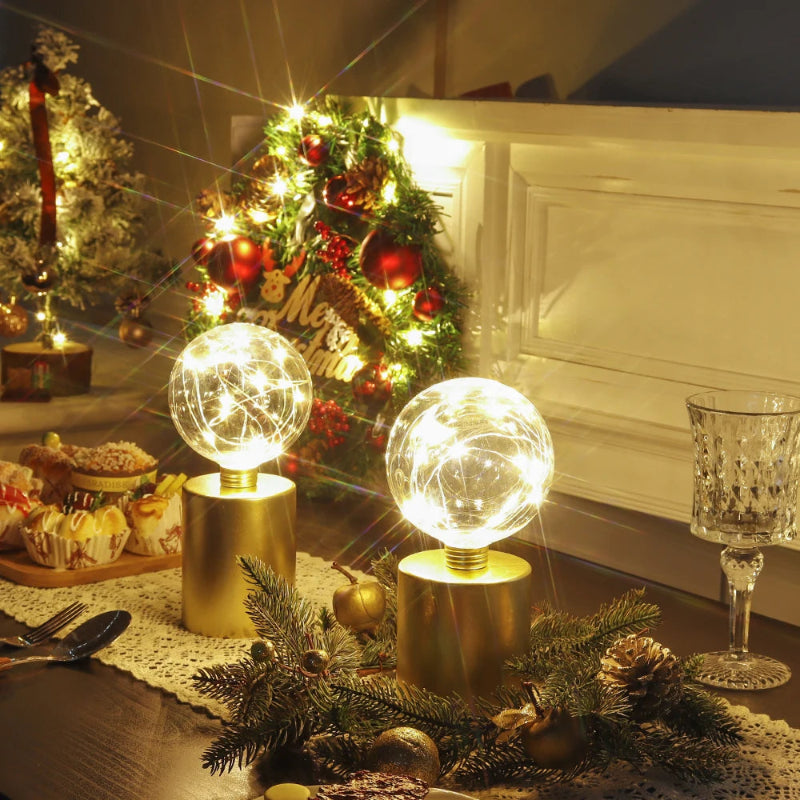 A holiday-themed table setting with two modern table lamps with gold cylindrical bases and clear glass globe shades filled with fairy lights, surrounded by festive decorations, a glass, and food.
