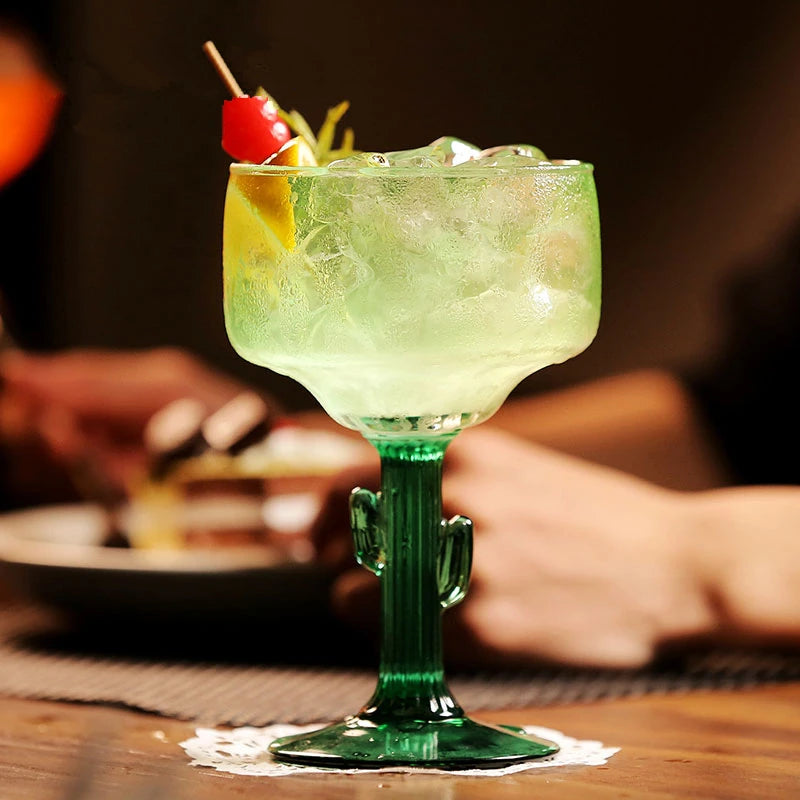 Green cocktail in a cactus-shaped glass with a cherry on a wooden table.