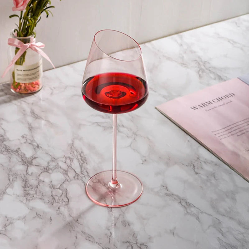 A wine glass with a pink stem and an angled rim, filled with red wine, on a marble surface.