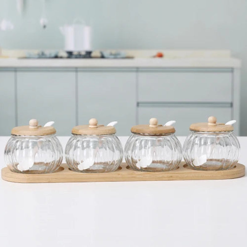 Four empty pumpkin-shaped glass jars with wooden lids and spoons, on a bamboo tray.