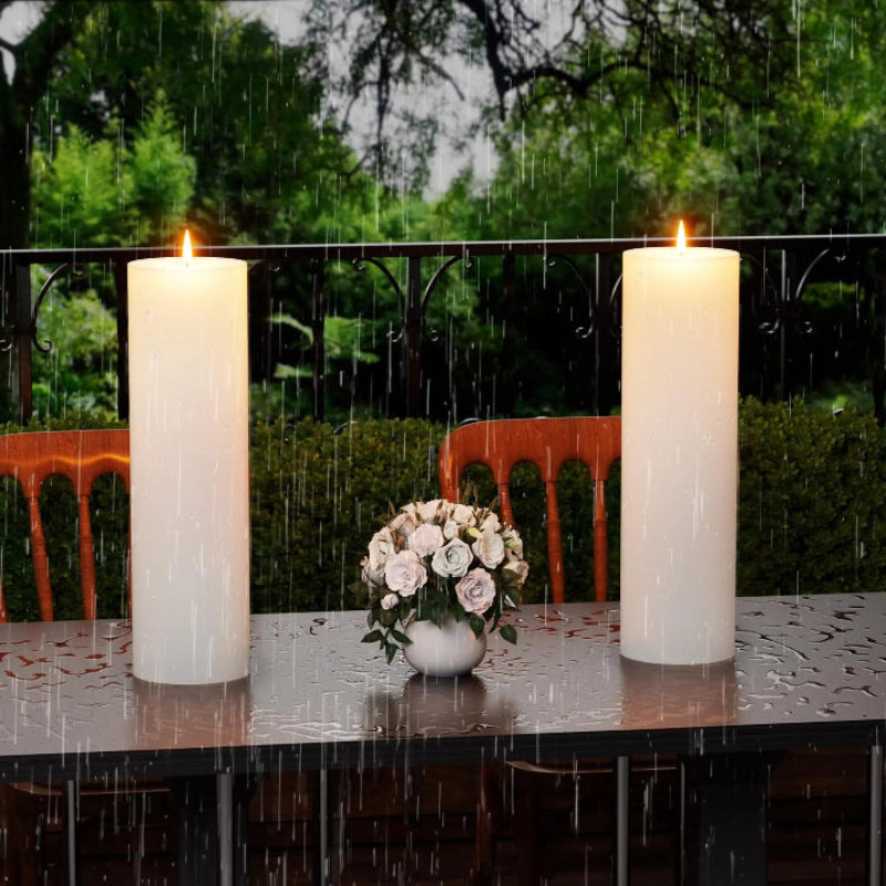 Two flameless outdoor candles and a small bouquet of roses sit on a wet table in the rain, with a green garden in the background.