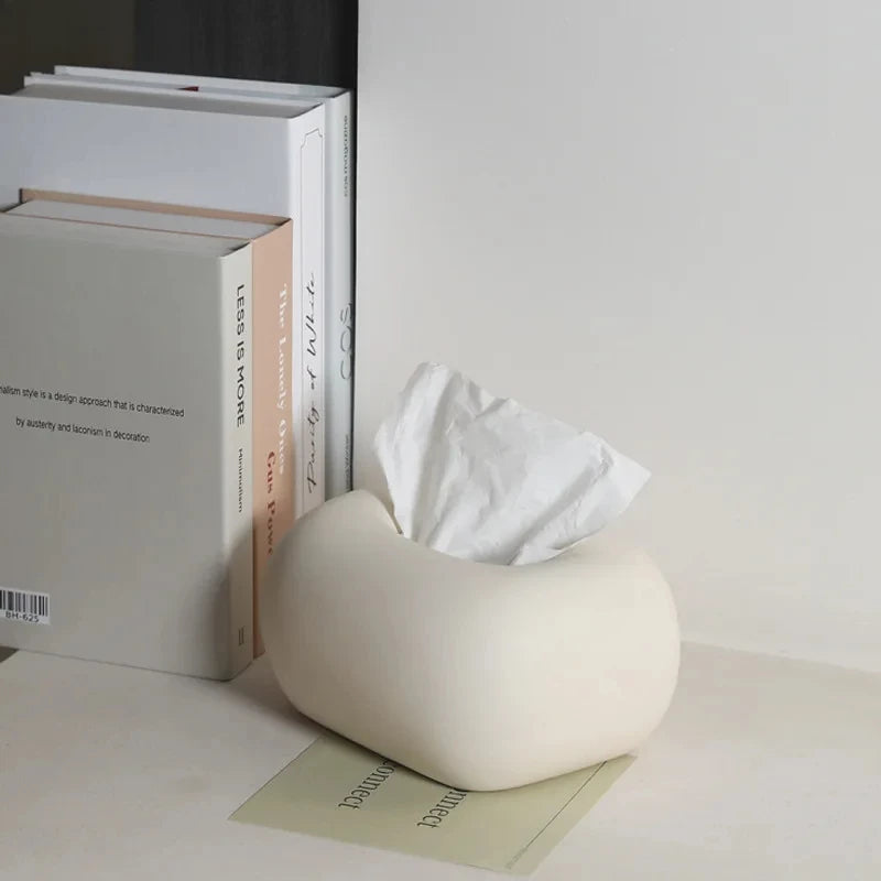 A white, donut-shaped tissue box holder sits on a light-colored surface with a stack of books behind it.