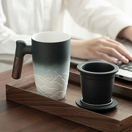A stylish ceramic thermal mug with a wooden handle, featuring an elegant mountain design, placed on a wooden tray. A matching black tea strainer sits beside it. In the background, a person in a white shirt is typing on a laptop.