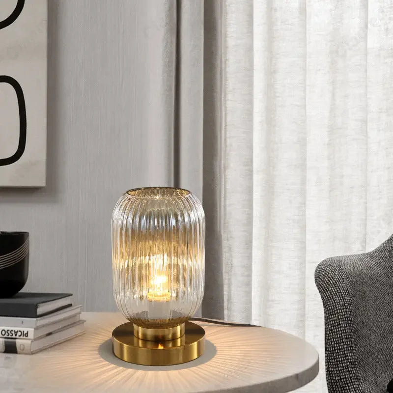 Lit amber glass lamp with a gold base on a round table next to books and a chair.