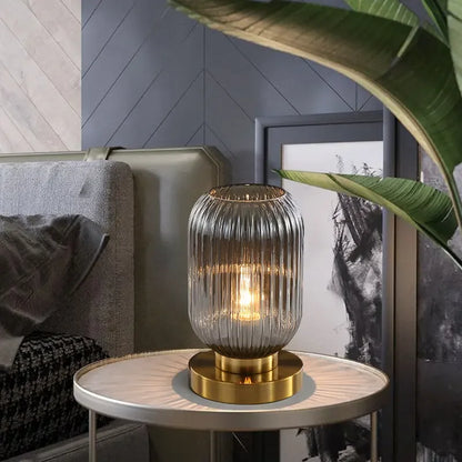 A lit, ribbed, smoky gray glass table lamp with a gold base sits on a small round table next to a bed and framed artwork. Part of a large green plant is visible in the foreground.