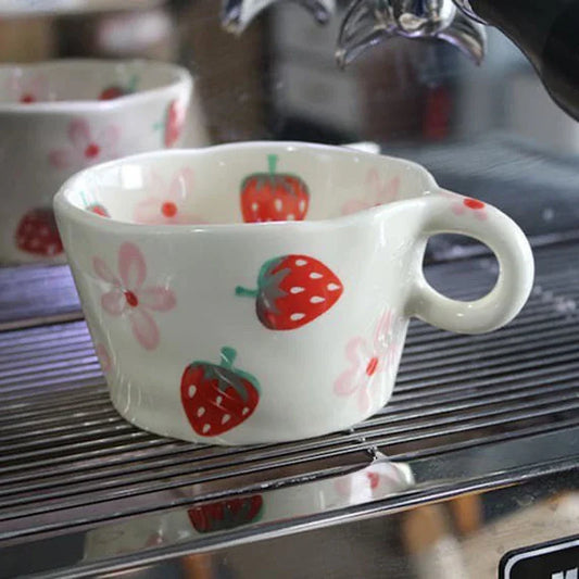 A close-up shot of a white ceramic mug with a rounded handle, featuring hand-painted red strawberries and pink flowers. The mug is placed on a stainless steel grate with parallel lines, suggesting a commercial or industrial setting, possibly part of a coffee machine or kitchen equipment. Another mug with a similar design is partially visible in the blurred background. The focus is on the intricate details of the strawberry pattern on the foreground mug.