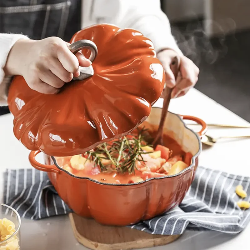 A person lifts the pumpkin-shaped lid off an orange cast iron pot filled with a steaming stew.