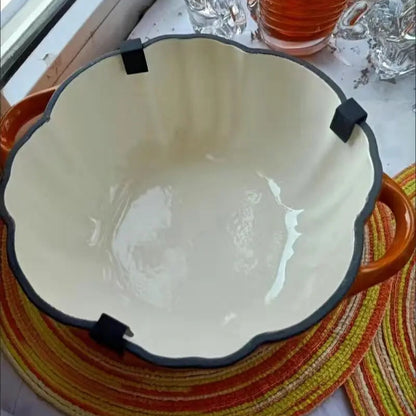The view from above shows a large, round, off-white bowl with a scalloped, dark-rimmed edge and two orange handles, sitting on a striped placemat.