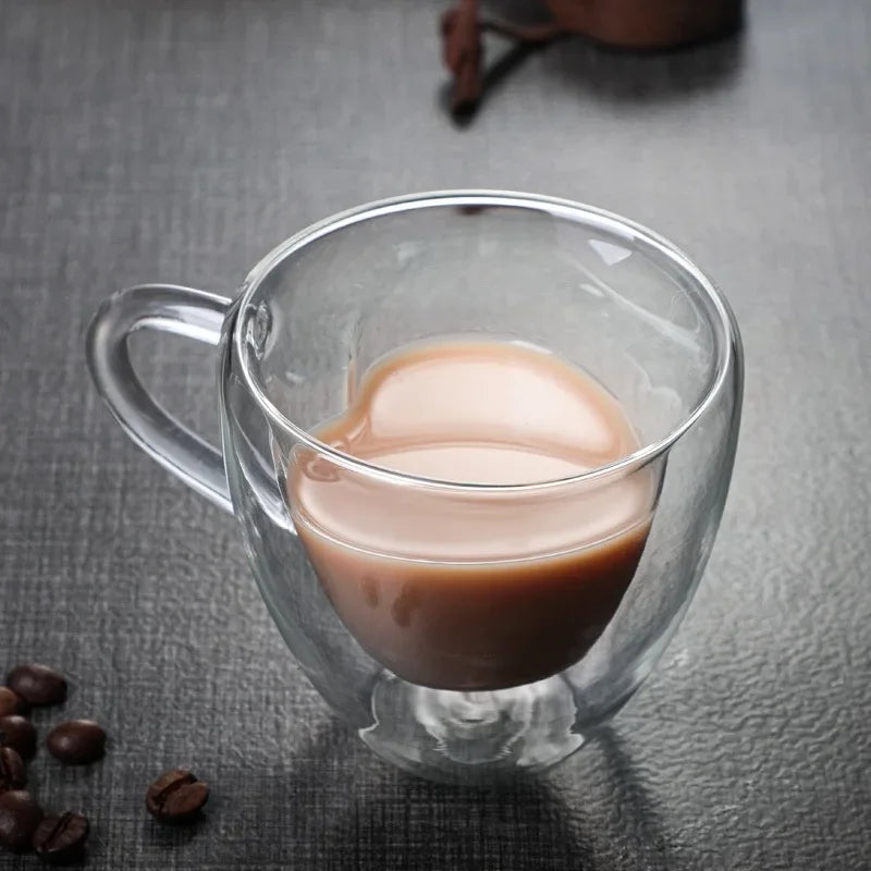 A clear, heart-shaped glass mug containing coffee is on a dark surface, with coffee beans scattered nearby.
