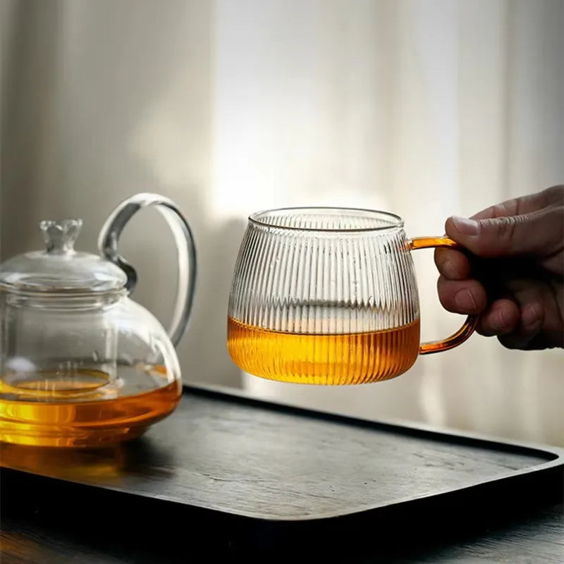 A hand holding a ribbed glass mug with an amber-colored handle, filled with a gold liquid. A clear teapot is on a tray to the left.