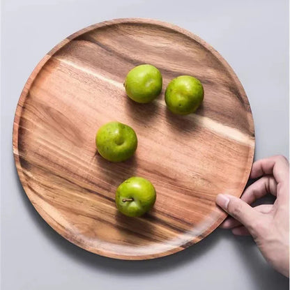 A hand holds a round wooden plate with four green plums on it.