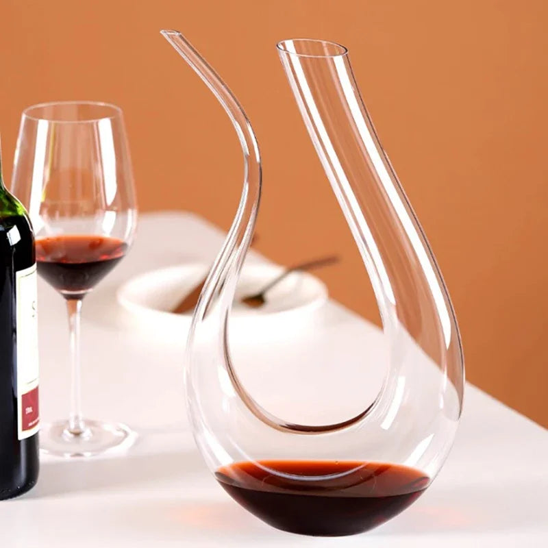 A swan-shaped glass wine decanter with a small amount of red wine at the bottom, next to a wine glass and bottle.