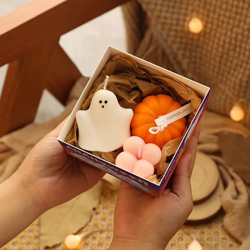 Two hands hold an open box containing three Halloween-themed candles nestled in brown crinkle-cut paper. The box has a colorful design along its edges. Inside are a white ghost-shaped candle with two black dot eyes, an orange pumpkin-shaped candle with a white string tied around it, and a light pink four-leaf clover-shaped candle. Soft, warm-toned string lights are blurred in the background. The hands appear to be light-skinned.