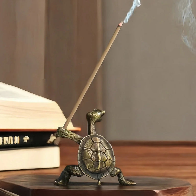 A lit incense stick held by a brass turtle figurine, with smoke curling upward, on a wooden surface next to books.