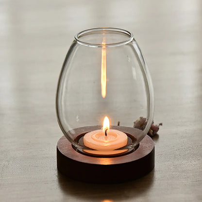 A minimalist glass candle holder with a wooden base, featuring a softly glowing tealight candle. The transparent glass creates a reflection of the flickering flame, adding warmth and elegance to the neutral-toned setting.