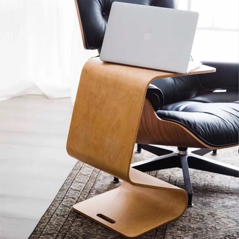 A bent plywood side table with a light wood finish, featuring a laptop on its surface, positioned next to a black leather Eames lounge chair on a patterned rug.