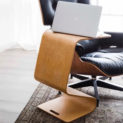 A bent plywood side table with a light wood finish, featuring a laptop on its surface, positioned next to a black leather Eames lounge chair on a patterned rug.