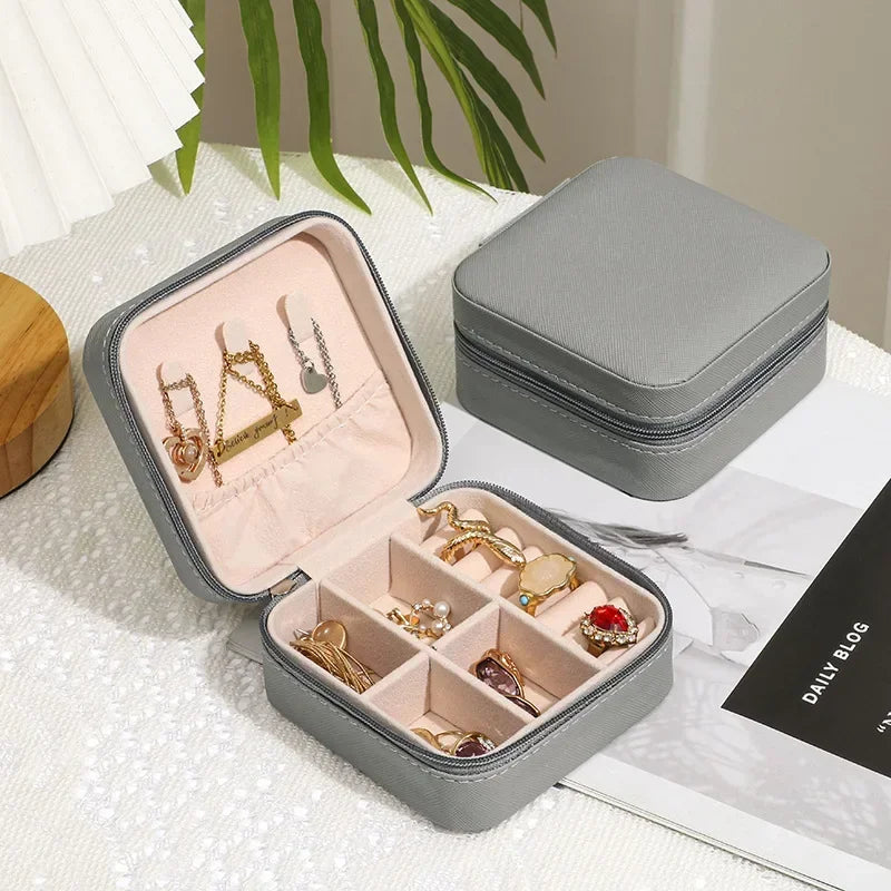 Two grey, square, zippered jewelry boxes, one open to display various rings and earrings in organized compartments.