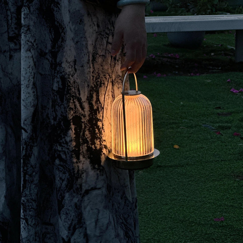 A person's hand holding a glowing portable lamp with a ribbed glass shade outdoors at dusk.