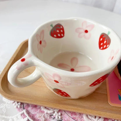 A close-up shot of a white ceramic mug with hand-painted red strawberries and pink flowers, resting on a square light-colored wooden tray. The mug has a rounded handle and a slightly flared rim. A portion of a pink and white patterned cloth is visible beneath the tray, adding a touch of texture to the composition. The focus is on the mug's design and its placement on the tray.