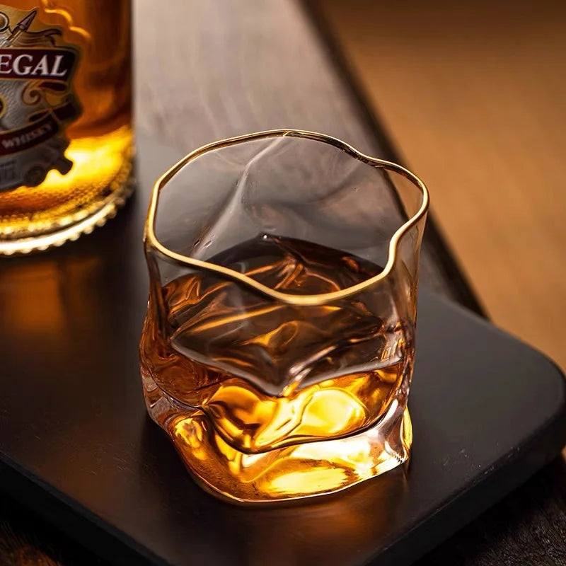 A glass with a crumpled texture and a gold rim, filled with a brown liquid and a single piece of ice, placed next to a bottle of Regal whisky.