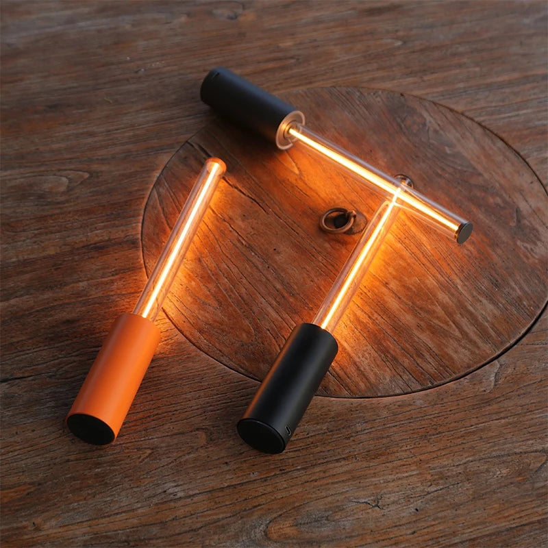 Two different colored cylindrical lamps with glowing tubes, one orange and one black, crossed on a round wooden table.