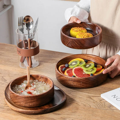 A set of walnut bowls displayed on a wooden table, filled with fresh fruit slices, soup, and pastries. A person is holding one bowl, showcasing its smooth, polished finish. A glass container with cutlery is placed in the background, adding a rustic dining ambiance.