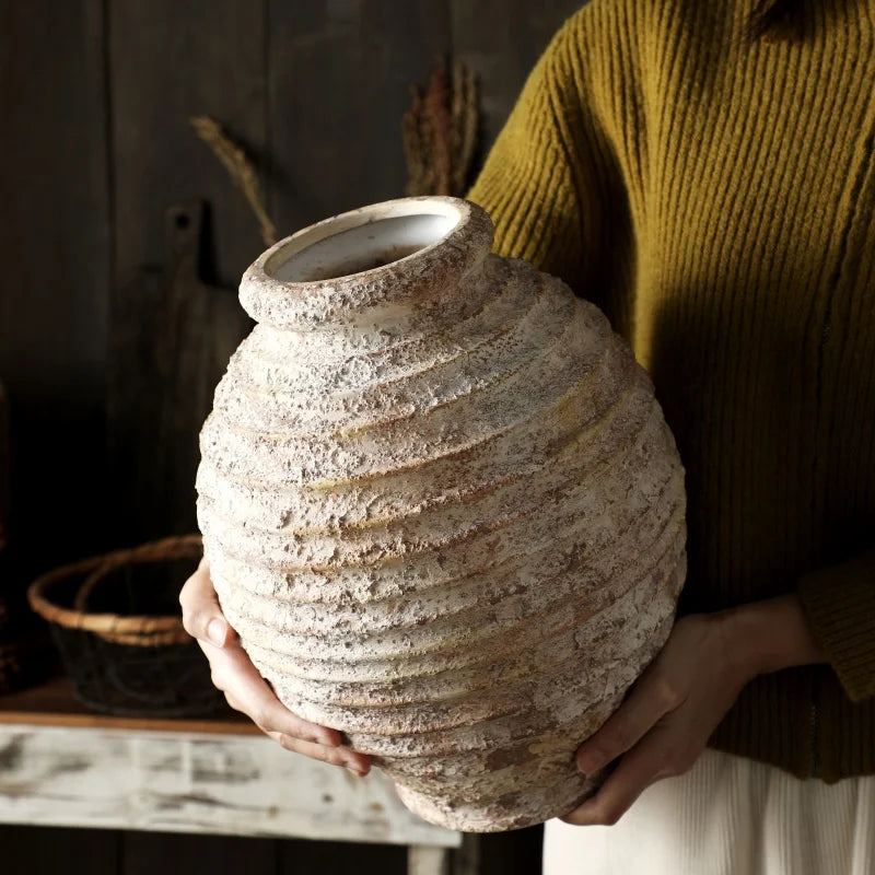 A person in a mustard-yellow sweater holds a large, round, heavily textured, rustic ceramic vase.