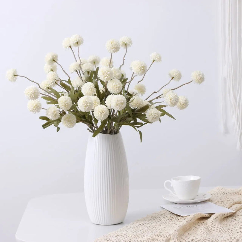 A ribbed white vase holds a bouquet of white artificial dandelion-like flowers, next to a teacup and a lace cloth.