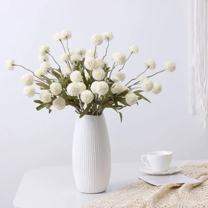 A ribbed white vase holds a bouquet of white artificial dandelion-like flowers, next to a teacup and a lace cloth.