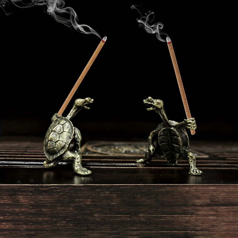 Two detailed bronze incense holders shaped like turtles holding burning incense sticks, with smoke rising from them, against a black background.