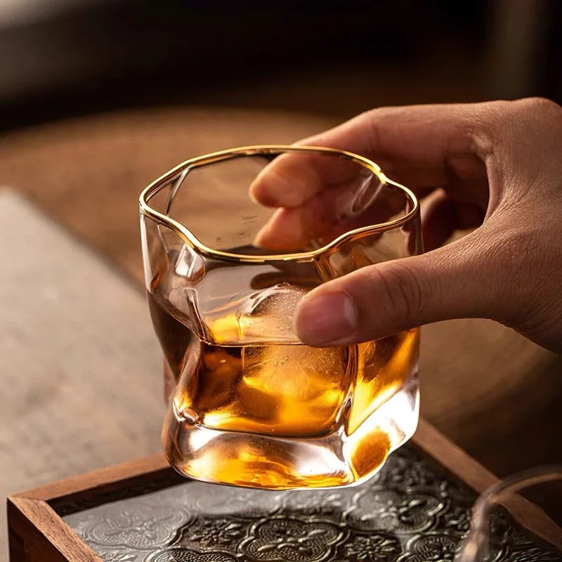 A hand holding a unique, crumpled-looking glass with a gold-rimmed edge, filled with a brown liquid and a piece of ice.