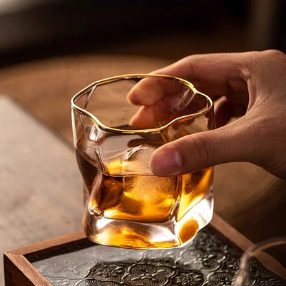 A hand holding a unique, crumpled-looking glass with a gold-rimmed edge, filled with a brown liquid and a piece of ice.