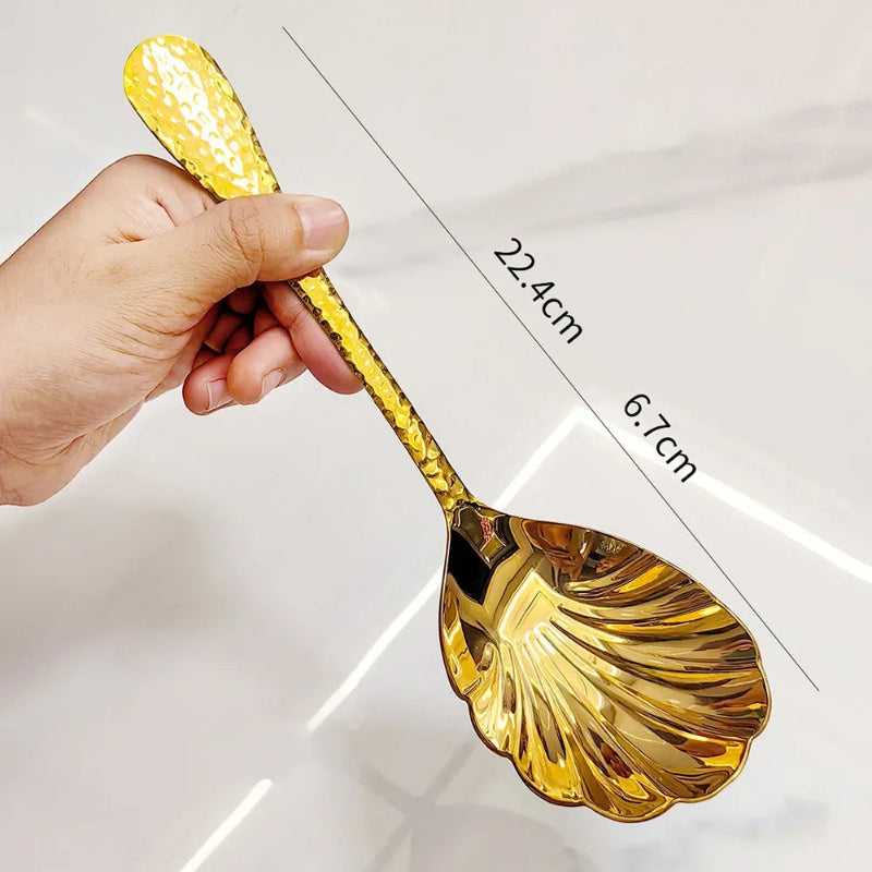 A hand holding a single gold serving spoon with a hammered handle and a scalloped, shell-shaped head. The image is labeled with its dimensions.