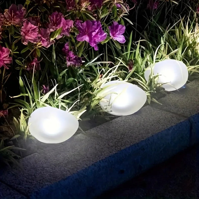 Three white, glowing, egg-shaped lights line the edge of a garden bed with grass and purple flowers.