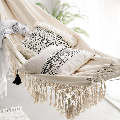 A beige fabric hammock with macrame fringe and a black tassel is adorned with two pillows featuring black geometric patterns and a striped throw blanket.