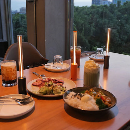 Three small, glowing, cylindrical lamps on a restaurant table, illuminating a meal and drinks.