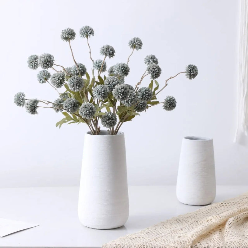 A white vase with light blue artificial flowers sits next to a smaller white vase and a cream-colored lace cloth.