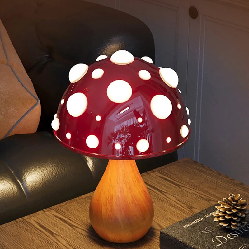 A mushroom-shaped lamp with a dark red cap and white spots, on a wooden table next to a book and a pinecone.