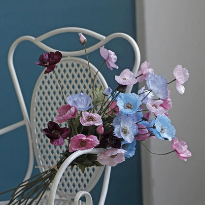 A bouquet of pink, blue, and purple poppies rests on a white wicker chair.