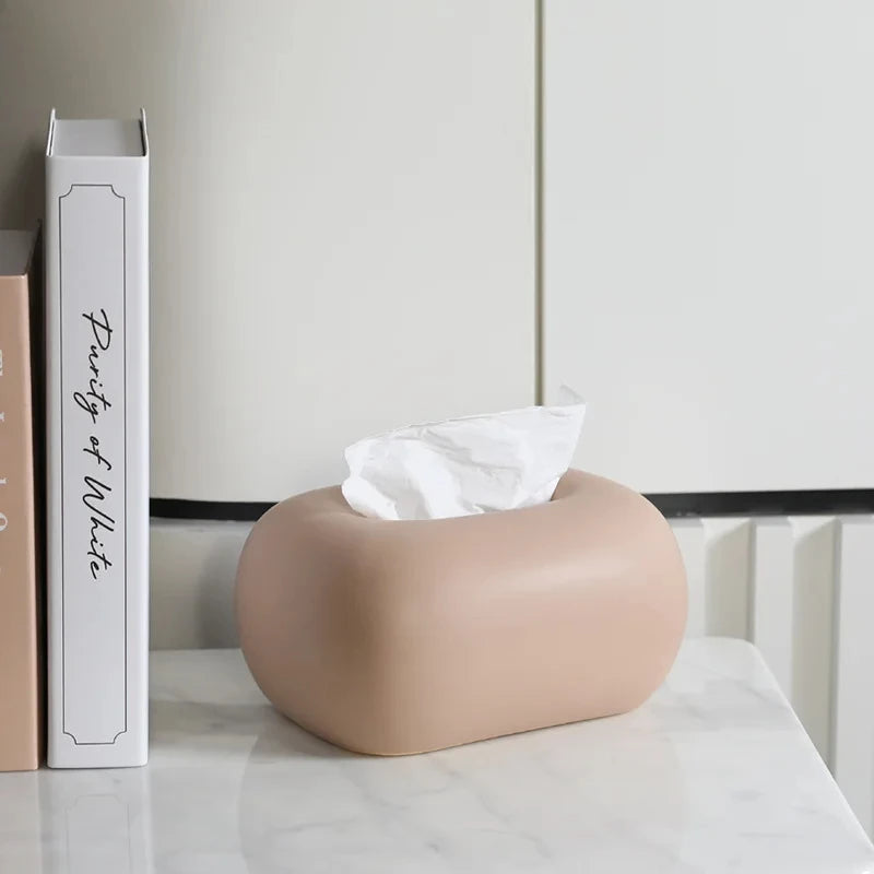 A donut-shaped, beige tissue box holder sits on a marble-patterned surface next to a stack of books.