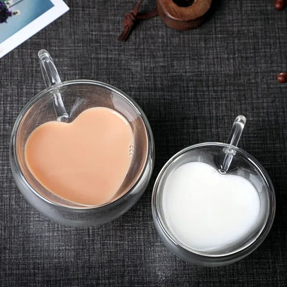 Two heart-shaped glass mugs are on a dark surface, one filled with coffee and the other with milk.
