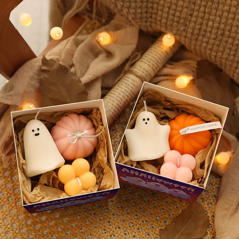 Two open boxes showcase Halloween-themed candles. The box on the left contains a white ghost-shaped candle with black dot eyes, a pink pumpkin-shaped candle with a string tied around it, and two stacked orange cube-shaped candles. The box on the right displays a white ghost-shaped candle with black dot eyes, an orange pumpkin-shaped candle, and a light pink four-leaf clover-shaped candle. The boxes are arranged on a textured surface with soft, warm-toned string lights scattered around them.
